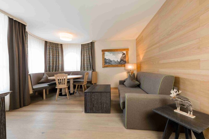 Ein gemütliches Wohnzimmer mit einer Eckcouch, einem hölzernen Couchtisch, einem Esstisch mit Stühlen und einer Bank, großen Fenstern mit Vorhängen und einem Wandbild von einer Berglandschaft.