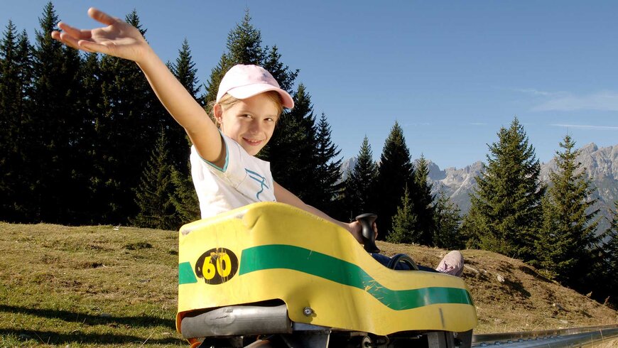 Ein Kind sitzt in einer Sommerrodelbahn und wirft beim Aktivurlaub in Tirol die Hände in die Luft. 