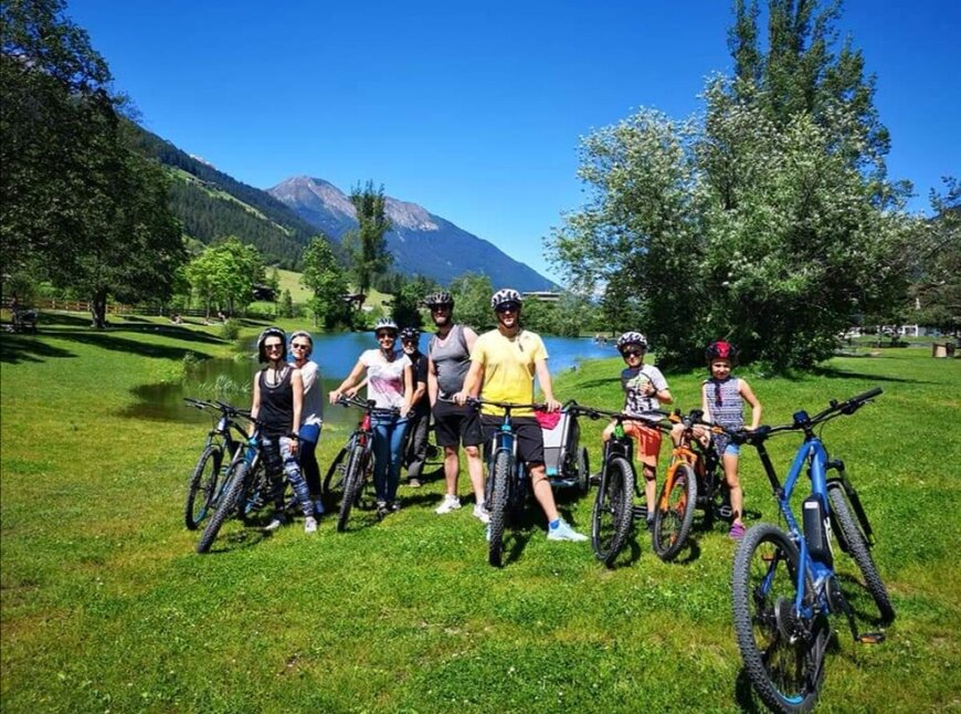 Mehrere Urlauber sind bei strahlendem Sonnenschein mit ihren Fahrrädern bei einem See unterwegs. 
