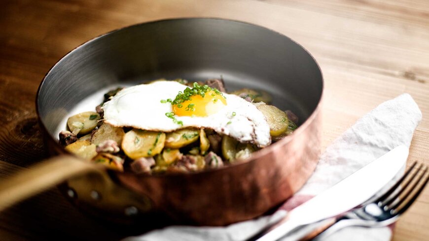 Ein Spiegelei mit Schnittlauch serviert auf Bratkartoffeln in einer kupferfarbenen Pfanne.