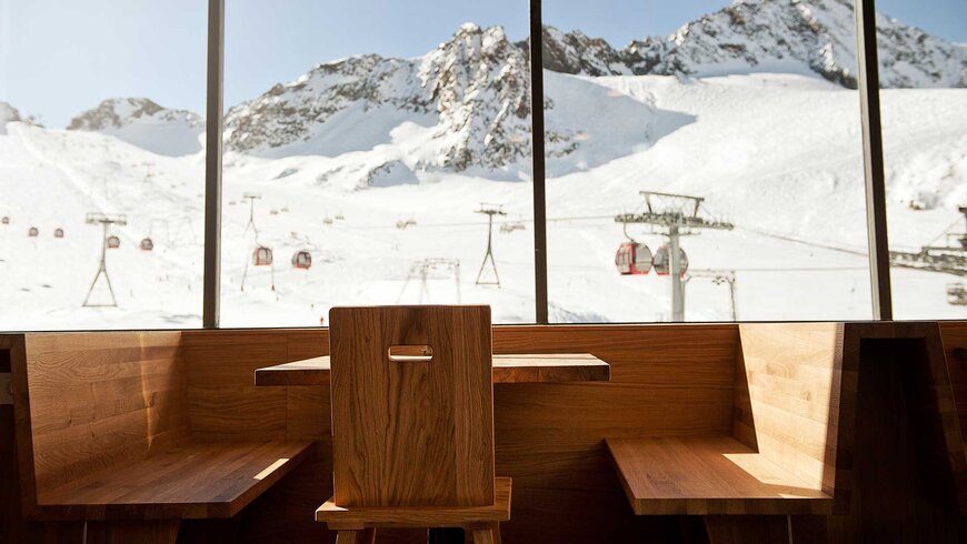 Blick von einem Tisch einer Berghütte hinaus auf die Gondelbahn vor verschneiten Bergen.