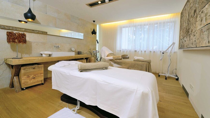 Spa-Behandlungsraum mit Massageliege, holzverkleidetem Interieur, großer Spiegelfront, dezenter Beleuchtung und entspannender Atmosphäre.