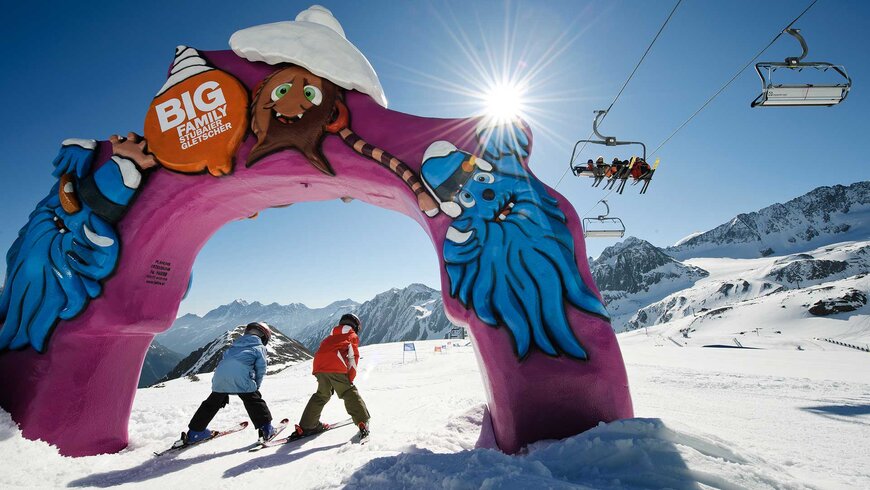 Zwei Kinder in Skiausrüstung fahren durch ein farbenfrohes Tor mit einem Comic-Murmeltier und Schneemann auf einem sonnigen Skigebiet mit Skilift im Hintergrund.