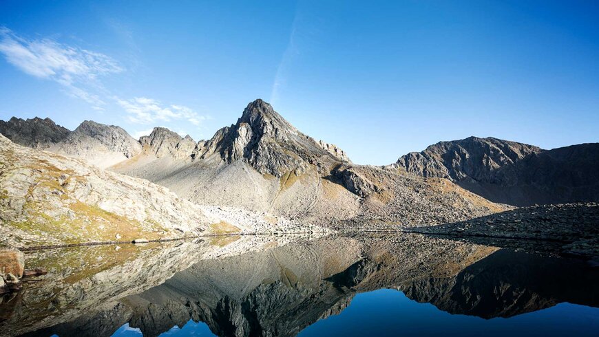 Spiegelung von scharfkantigen Berggipfeln in einem ruhigen Gebirgssee unter klarem blauen Himmel.