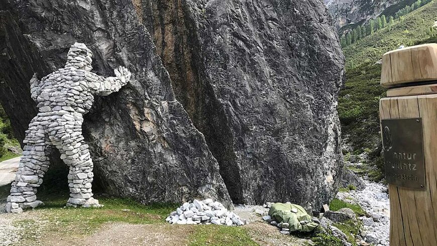 Steinfigur eines Menschen, die so positioniert ist, als würde sie gegen eine große Felswand in einer grünen Berglandschaft drücken, neben einem Schild mit der Aufschrift "Natur Kunzt".