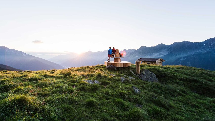 Zwei Wanderer genießen den Sonnenuntergang auf einer Aussichtsplattform in den Tiroler Alpen während ihres Sommerurlaubs.