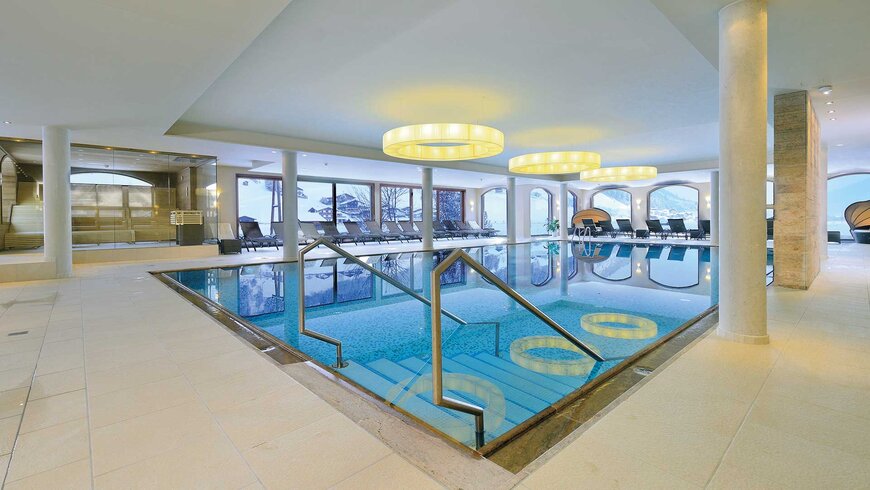 Modernes Hallenbad in einem Hotel mit Glasgeländer, Liegestühlen und auffälligen runden Deckenleuchten.