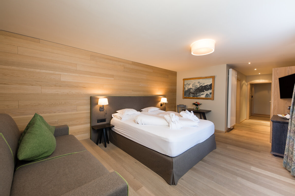 Hotelzimmer mit einem großen Doppelbett, Nachttischen mit Lampen, einer Sitzcouch, Parkettboden und einer Wand mit Holzverkleidung sowie einem Bild von einem Bergpanorama.