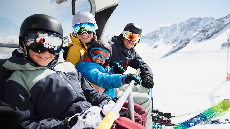 Eine vierköpfige Familien sitzt mit Skikleidung im Lift und lächelt in die Kamera.