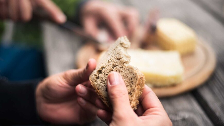 Eine Person hält ein Stück frisches Brot in den Händen, während im Hintergrund Käse auf einem Holzbrett liegt.