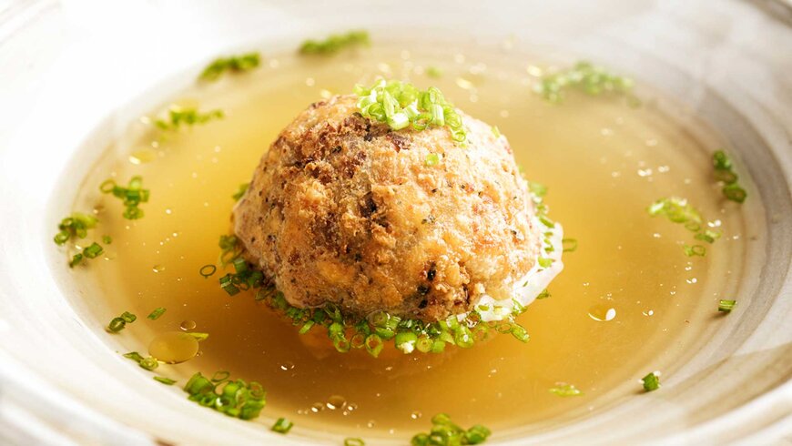 Nahaufnahme einer traditionellen Tiroler Speckknödelsuppe, serviert in einer klaren Brühe und garniert mit frischen Schnittlauchringen.
