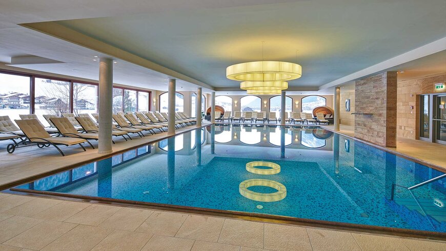 Luxuriöser Innenpoolbereich eines Hotels in Tirol mit Liegestühlen und großen Fenstern, die einen Blick auf die winterliche Landschaft bieten.