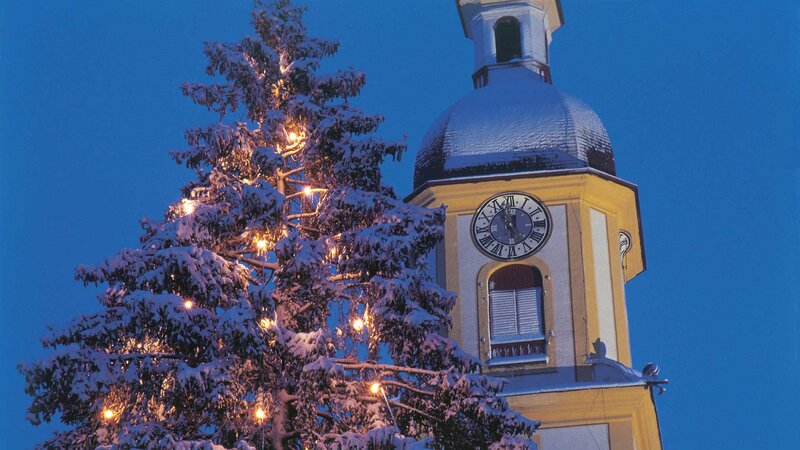 Schneebedeckter Kirchturm und festlich beleuchteter Weihnachtsbaum in Tirol bei Abenddämmerung.