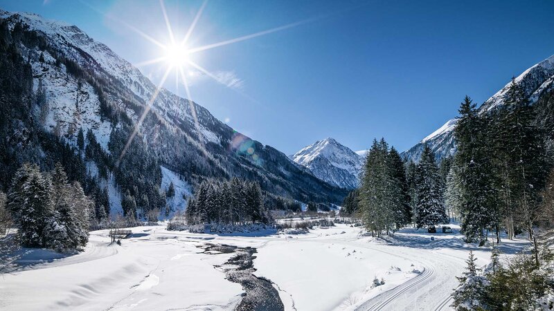 Sonnige Winterlandschaft in Tirol mit verschneiten Bergen, einem zugefrorenen Fluss und schneebedeckten Wäldern unter klarem, blauem Himmel.