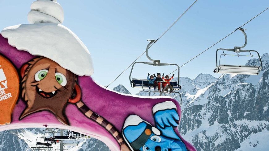 Bunte Kinderzone im Skigebiet Tirol mit fröhlichen Cartoon-Figuren, während ein Sessellift Skifahrer über die schneebedeckten Berge transportiert.