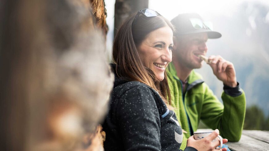 Lachende Frau und Mann in Outdoor-Kleidung genießen eine Pause mit Kaffee und Snack in den Bergen.