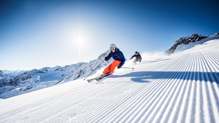 Zwei Skifahrer in farbenfroher Ausrüstung fahren eine frisch präparierte Piste hinunter bei strahlendem Sonnenschein und klarblauem Himmel.
