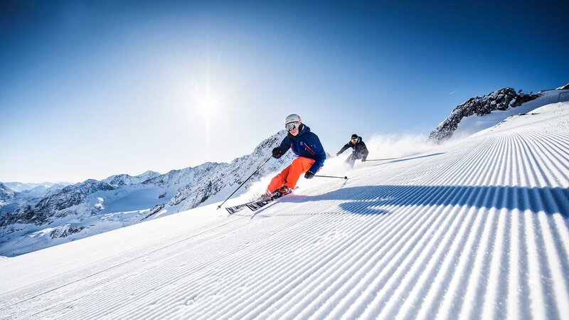 Zwei Skifahrer in farbenfroher Ausrüstung fahren eine frisch präparierte Piste hinunter bei strahlendem Sonnenschein und klarblauem Himmel.