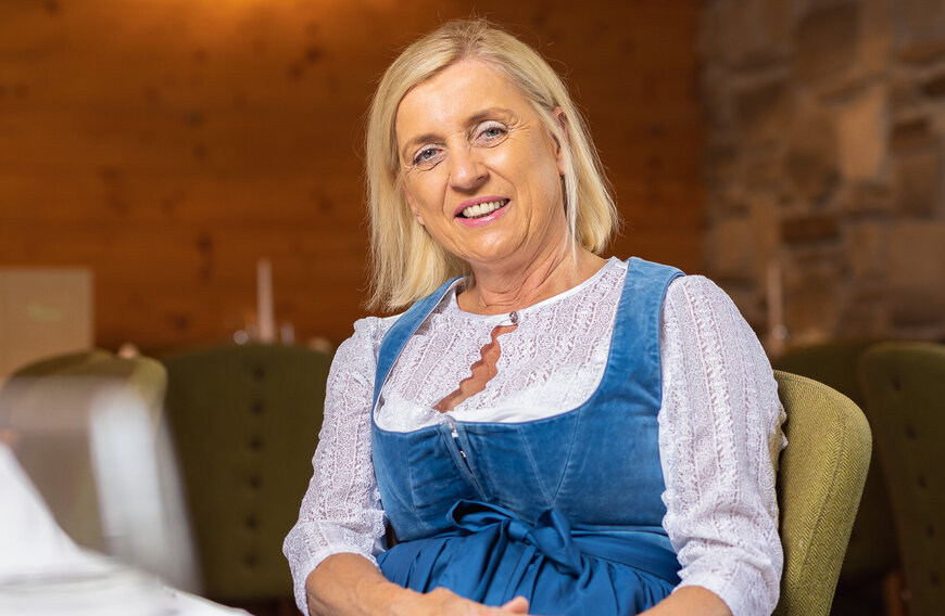 Eine blonde Frau in traditioneller Tiroler Tracht lächelt in einem gemütlichen Restaurant mit Holzverkleidung und gedämpfter Beleuchtung.