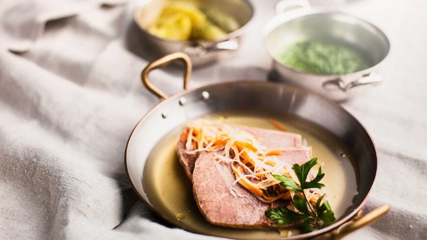 Traditionelles Tiroler Gericht mit zwei Scheiben gekochtem Fleisch, garniert mit Gemüse und Kräutern, serviert in einer Kupferpfanne, mit Beilagen in Schüsseln im Hintergrund.