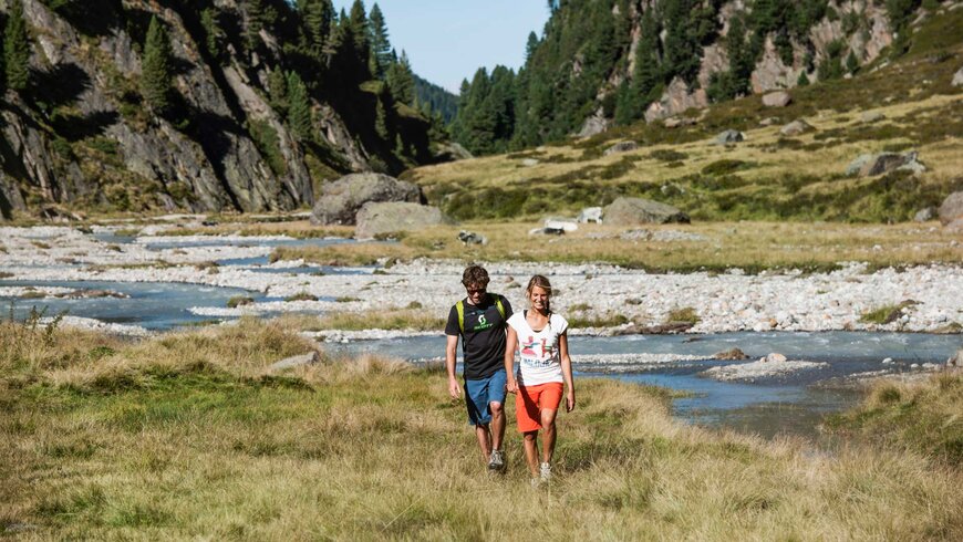 Ein Paar in sportlicher Kleidung wandert entlang eines Flusses durch eine malerische Berglandschaft mit grünen Wiesen und bewaldeten Hügeln.