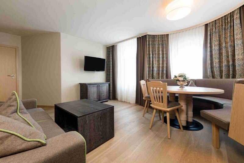 Helles, gemütliches Hotelzimmer mit Holzfußboden, einem Sofa, einem Flachbildfernseher, einem Holztisch und Stühlen, sowie großen Fenstern mit Vorhängen.