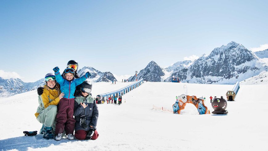 Glückliche Familie in Skikleidung posiert auf einer verschneiten Skipiste vor einer Kinderzone, umgeben von schneebedeckten Bergen und einem klaren blauen Himmel.