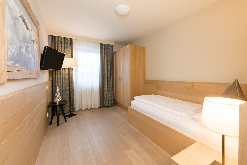Helles und gemütliches Einzelzimmer mit einem Einzelbett, Holzmöbeln, einem Flachbildfernseher an der Wand und großen Fenstern mit Vorhängen, die viel Tageslicht hereinlassen.