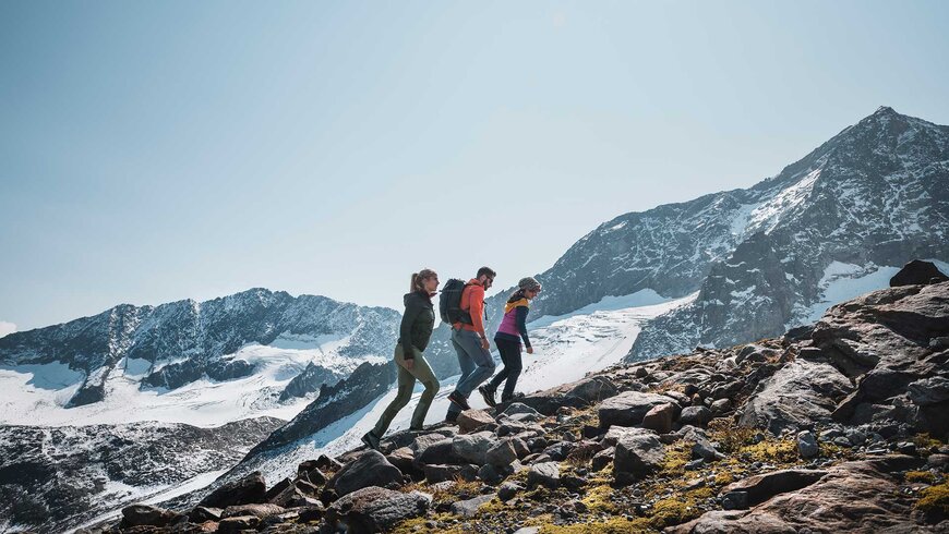 Drei Wanderer in sportlicher Kleidung erklimmen einen felsigen Bergpfad, mit schneebedeckten Gipfeln im Hintergrund unter einem klaren blauen Himmel.