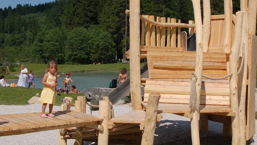 Ein kleines Mädchen in einem gelben Kleid spielt auf einem Holzspielplatz in der Nähe eines Sees, umgeben von grünen Bäumen und Wiesen.