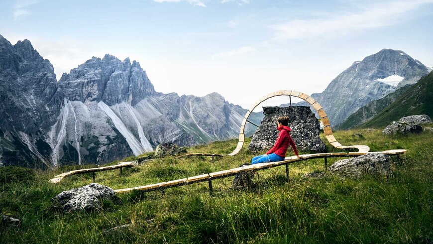 Person in roter Jacke sitzt auf einer kunstvollen Holzbank in einer Bergwiese, umgeben von steilen Felsformationen und grünen Hängen unter bewölktem Himmel.