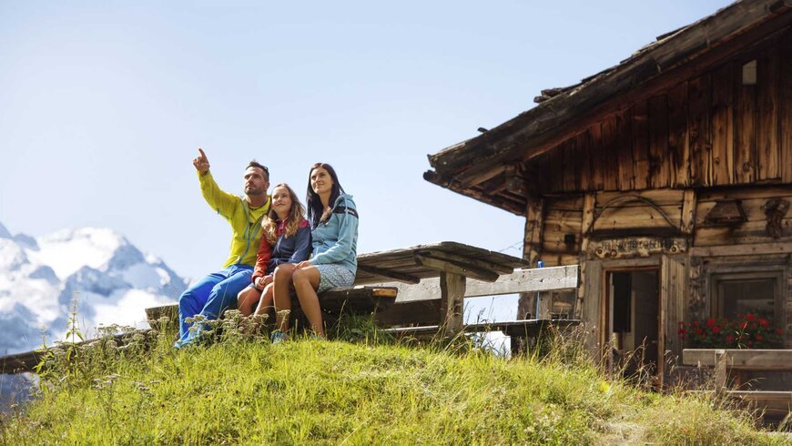 Eine Familie sitzt während dem Sommerurlaub in Tirol auf einer Bank neben dem rustikalen Berghotel im Stubaital und genießt den Ausblick auf die Berge im Hintergrund.