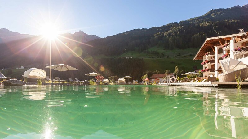 Der smaragdgrüne Naturbadeteich bei strahlendem Sonnenschein und dem Hotelgebäude direkt nebenan