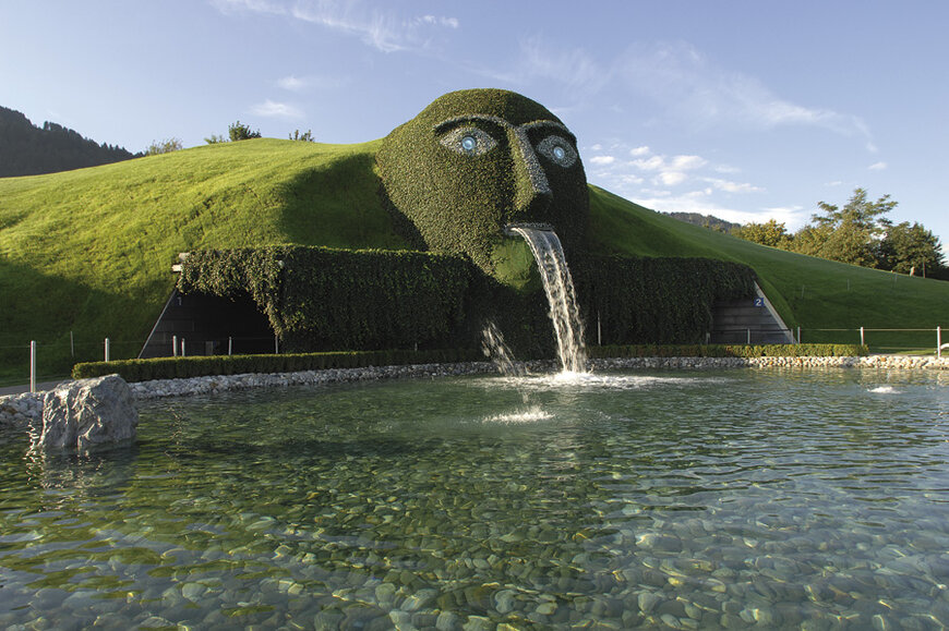 Große, mit Gras und Efeu bedeckte Skulptur eines Kopfes, aus dessen Mund Wasser in einen klaren Teich fließt, umgeben von grünen Hügeln und Bäumen.