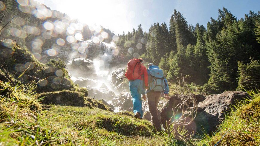 Zwei Wanderer mit Rucksäcken steigen im Wanderurlaub in Tirol einen sonnigen Waldpfad hinauf, in der Nähe eines rauschenden Wasserfalls.