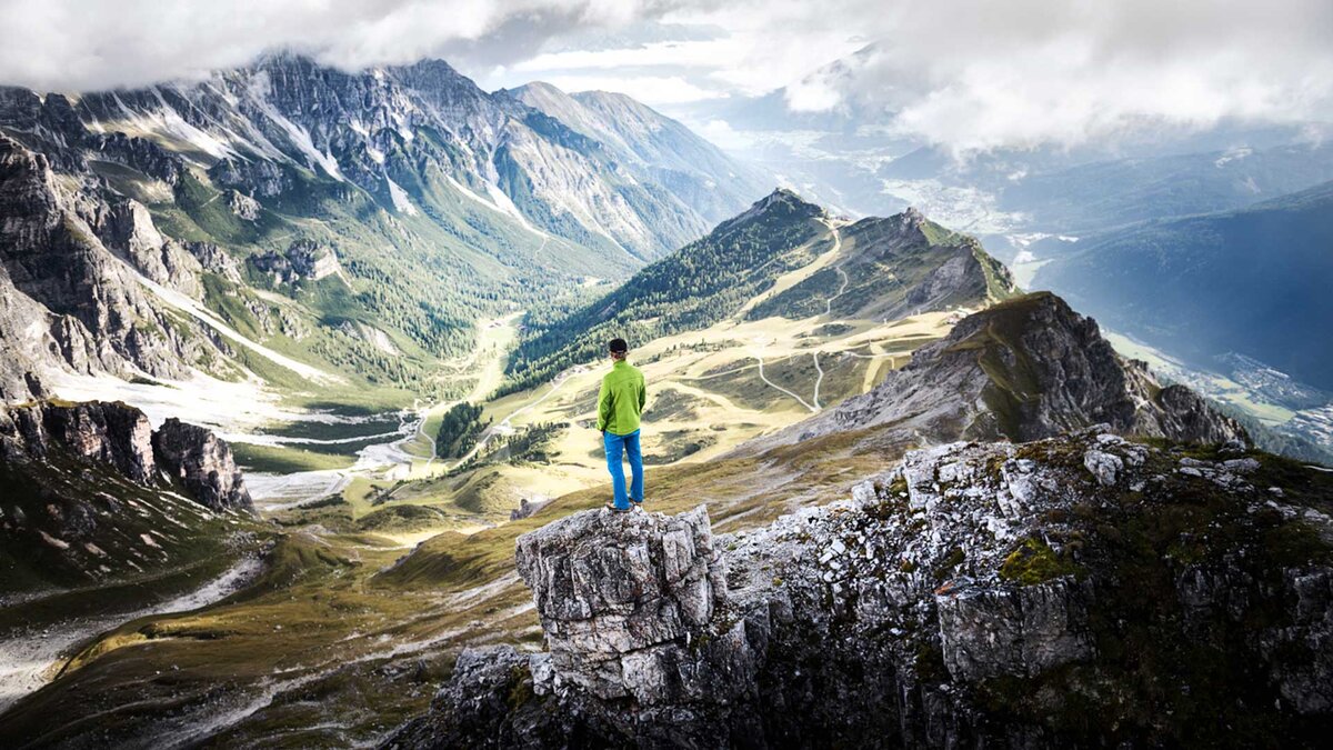Ein Wanderer in grüner Jacke steht auf einem Felsen und blickt auf ein weites, beeindruckendes Bergpanorama mit grünen Tälern und felsigen Gipfeln.