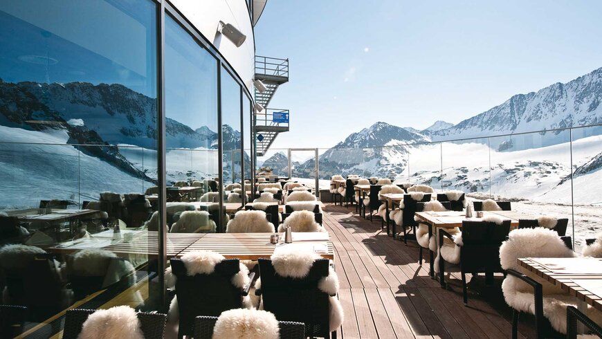 Sonnige Terrasse eines Bergrestaurants mit Tischen und Stühlen, die mit kuscheligen Fellen bedeckt sind, bietet einen atemberaubenden Blick auf schneebedeckte Berge.