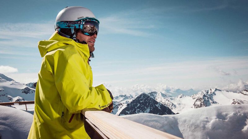 Ein Skifahrer im Aktivurlaub in Tirol in einer gelben Jacke und weißem Helm lehnt sich über ein Geländer und genießt die Aussicht auf schneebedeckte Berge und blauen Himmel.
