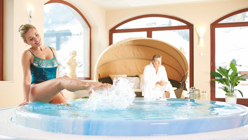 Eine Frau im Badeanzug sitzt Beim Wellness im Stubaital am Rand eines runden Whirlpools und spritzt spielerisch mit dem Wasser, während ein Mann in einem weißen Bademantel auf einer Liege im Hintergrund sitzt.