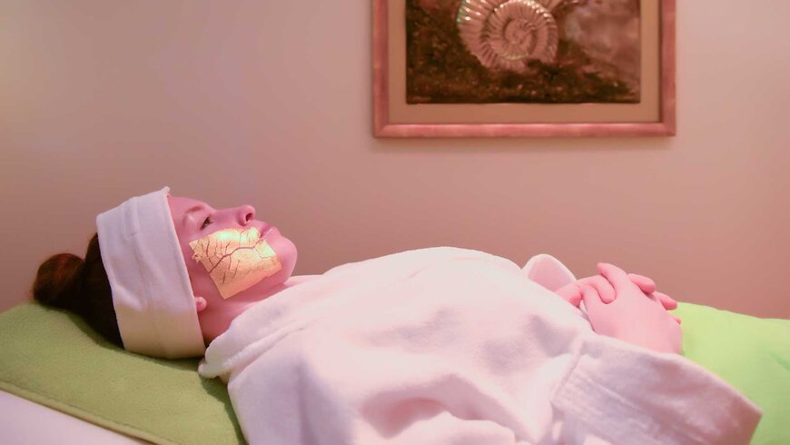 Eine Frau liegt entspannt auf einer Liege in einem Spa, während sie eine Gesichtsmaske mit goldenen Blättern trägt und einen weißen Bademantel und ein Stirnband trägt.