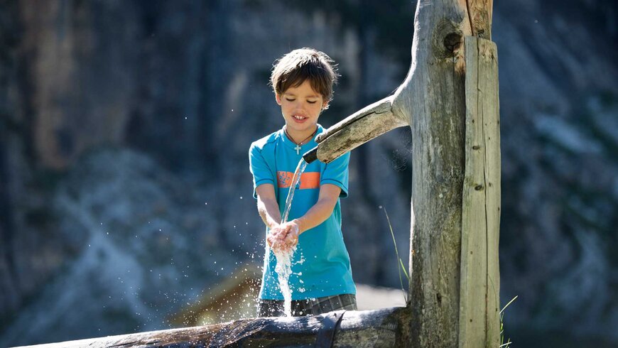 Ein Junge macht Sommerurlaub mit seiner Famlien in Tirol und spielt und erfrischt sich an einem rustikalen Holzbrunnen in den Bergen.