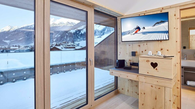 Ein Ausschnitt eines gemütlich in Holz gekleideten Hotelzimmers mit TV und großem Balkon mit Blick auf das Stubaital