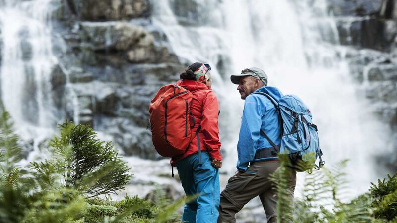 Zwei Wanderer mit Rucksäcken stehen während ihres Wanderurlaubs in Tirol vor einem rauschenden Wasserfall.