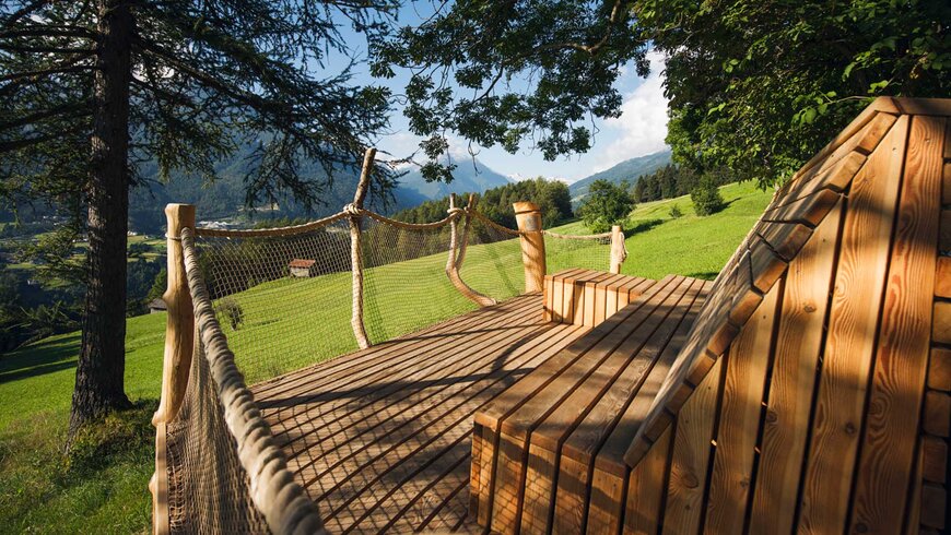 Holzplattform mit Sitzbank und Geländer aus Seilen, umgeben von grünen Wiesen und Bäumen, mit Blick auf die Tiroler Alpen im Sommerurlaub.
