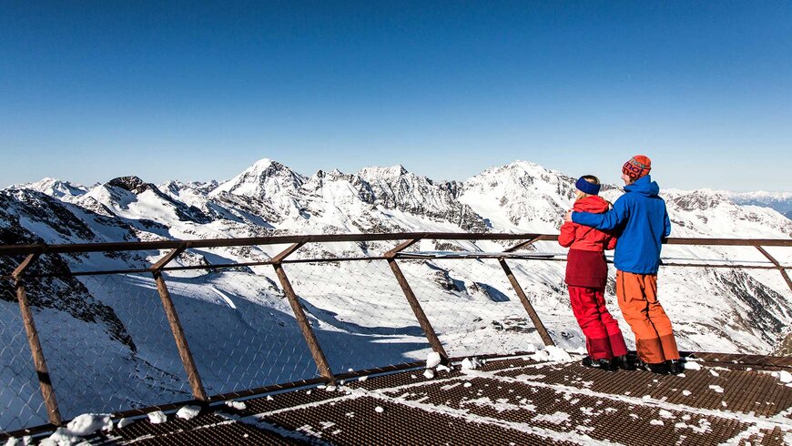 Ein Paar in bunter Winterkleidung genießt die Aussicht auf die verschneiten Alpen von einer Aussichtsplattform in Tirol.
