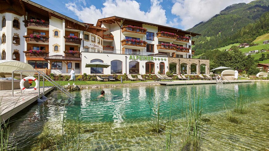 Ein im Naturbadeteich schwimmender Hotelgast mit dem Hotelgebäude im Hintergrund