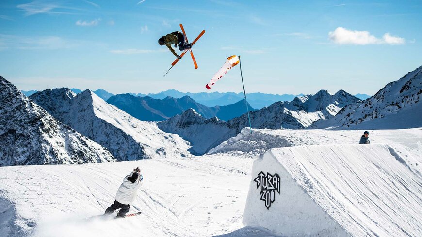 Ein Skifahrer springt über eine Schanze während der andere die Piste herunterfährt.
