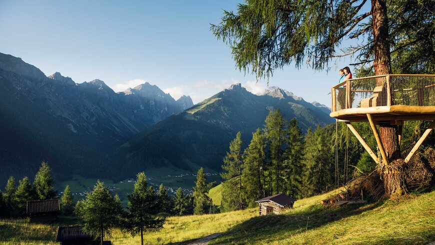Ein Paar genießt den Sommerurlaub in Tirol von einer Baumplattform aus, mit Blick auf eine malerische Berglandschaft im Sonnenschein.
