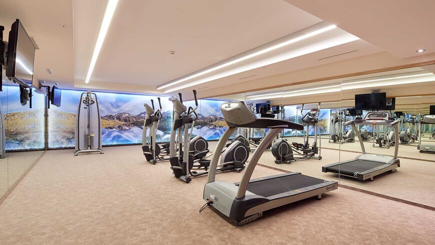 Ein moderner Fitnessraum mit Laufbändern, Crosstrainern und einem Bergpanorama im Hintergrund.