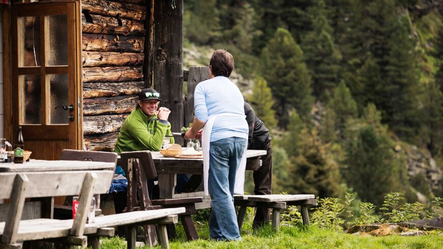 Eine Gruppe von Menschen sitzt an einem Holztisch vor einer rustikalen Berghütte.